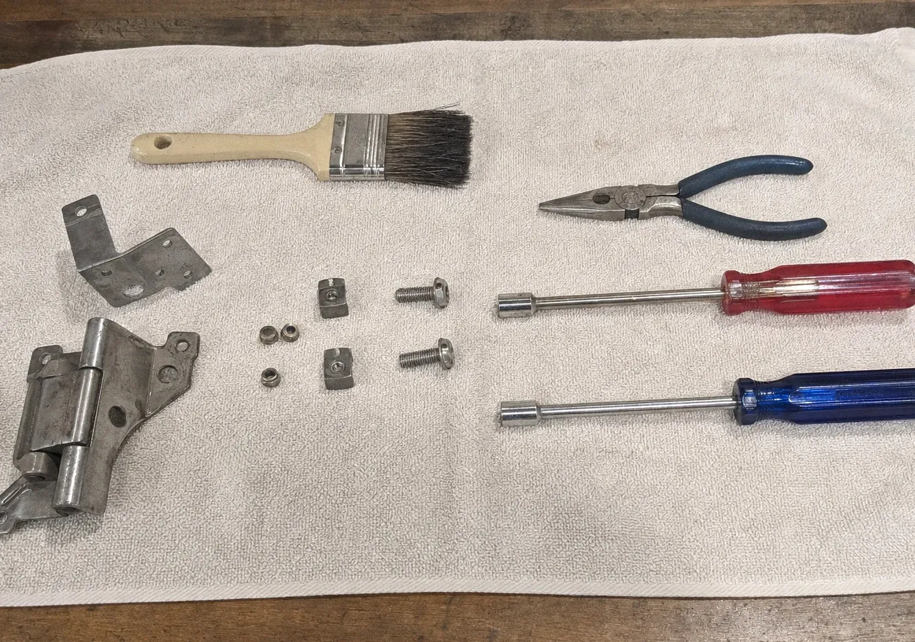Detail brush, needle-nose pliers, a nut driver, two screwdrivers and a hinge plate laid out on a white towel